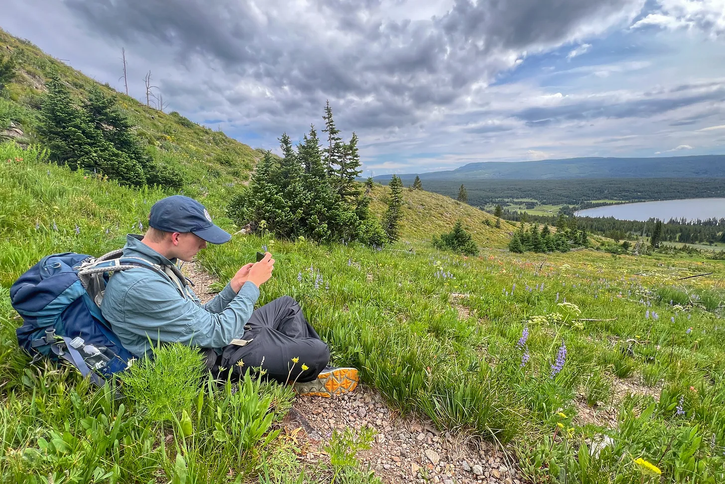 Young George sits on a grassy hillside in Yellowstone National Park, surrounded by wildflowers, with a backpack beside him. He looks at his phone with Heart Lake, pine trees, and dramatic clouds in the background.