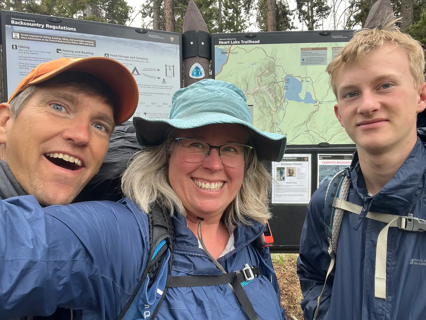 Jenny Golding, George Bumann, and young George smile in front of the Heart Lake Trailhead sign in Yellowstone National Park, ready for a backcountry hike. All three are wearing rain jackets and backpacks, with forest and trail maps visible in the background.