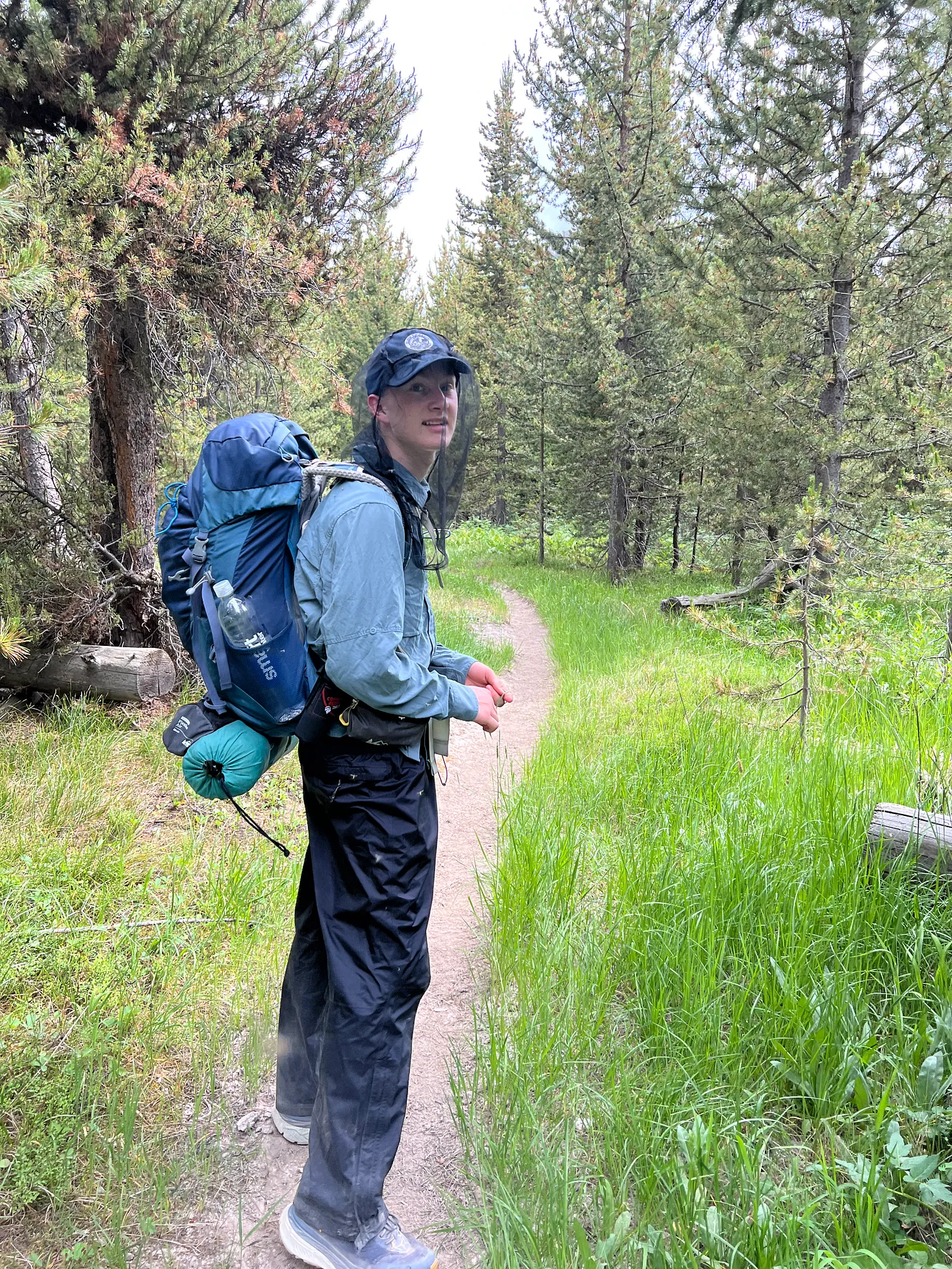 Young George stands on a forested trail in Yellowstone National Park, wearing a mosquito net hat and carrying a large backpack with camping gear. He looks back toward the camera, surrounded by tall pine trees and lush green grass.