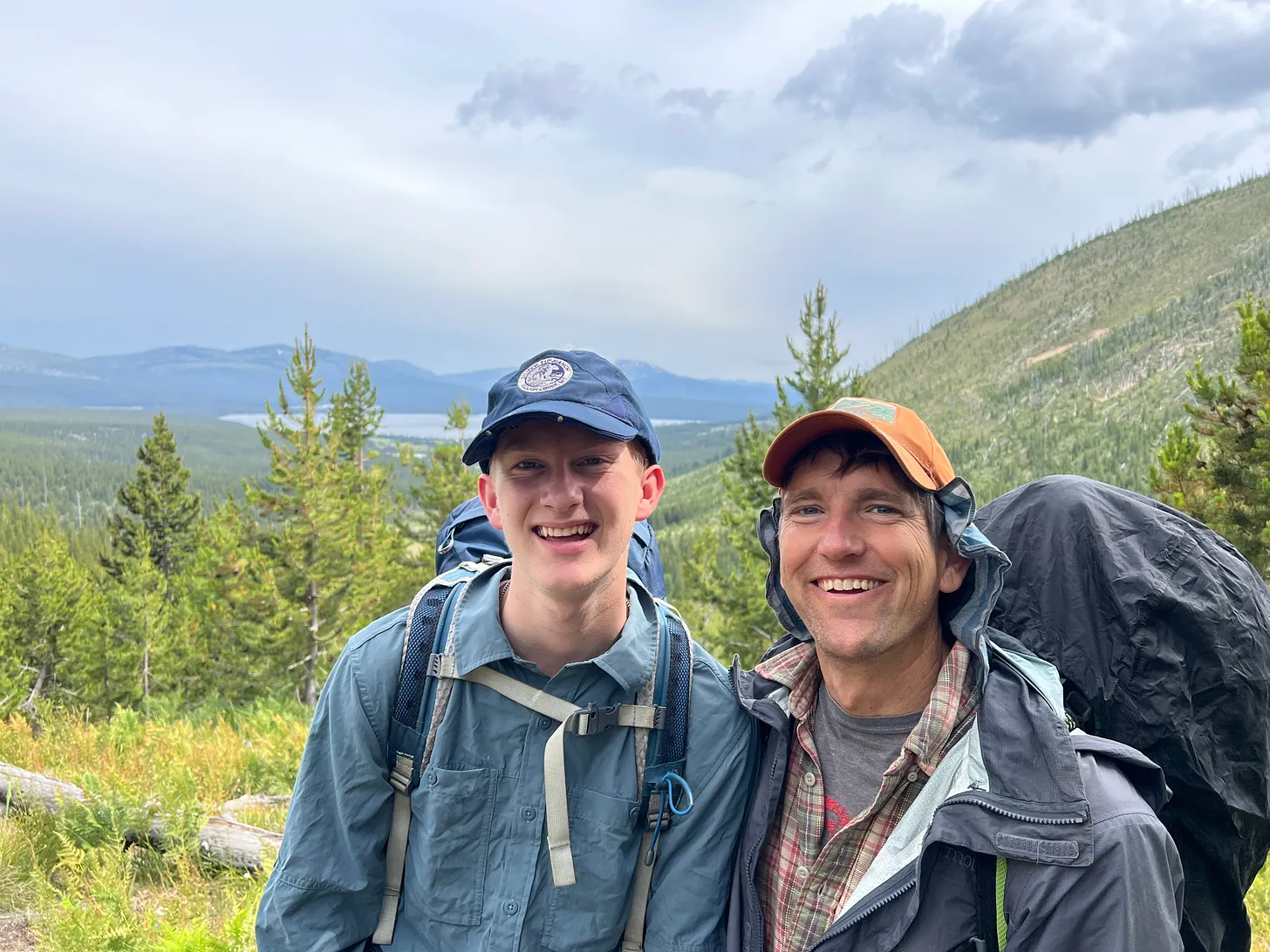 George Bumann and young George smile on a hiking trail in Yellowstone National Park, wearing backpacks and outdoor gear. Behind them are pine trees, a distant lake, and forested mountains under a cloudy sky.